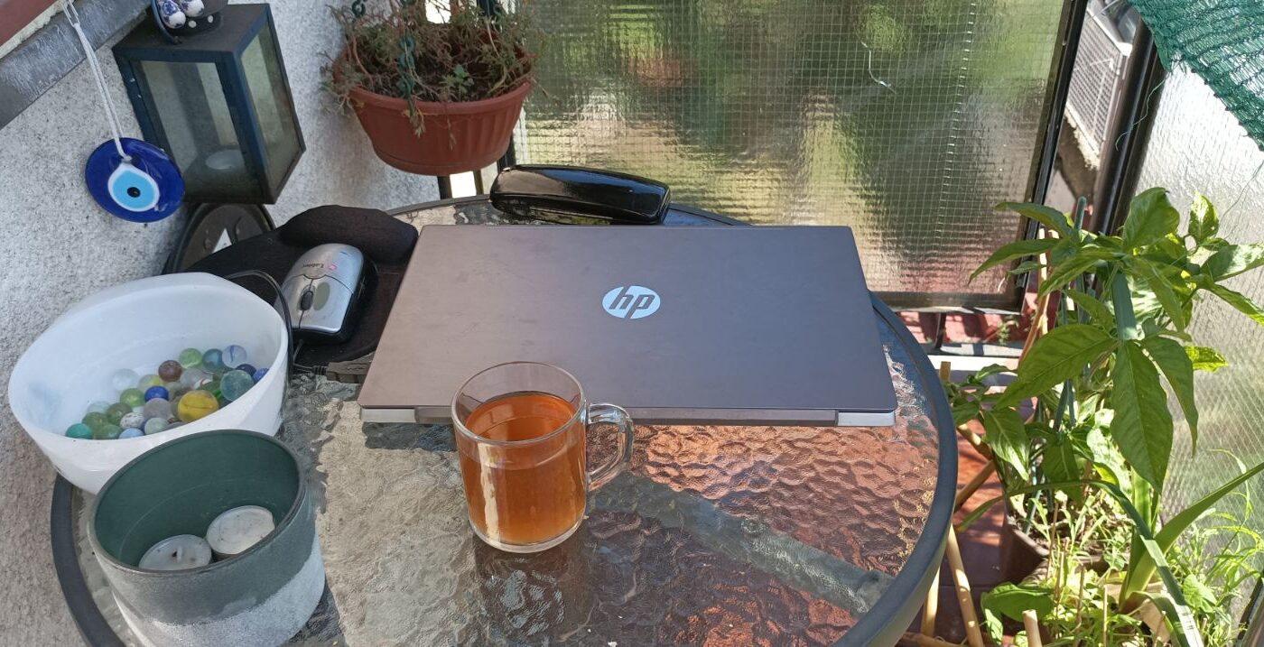 A cozy balcony workspace with a round glass table and potted plants on the right features a closed grey laptop and a mouse, a glass of ice tea, a white bowl of marbles, and a grey pot with tea candles. A blue lantern, a blue evil eye charm and a round pot with ground cover hang on the left.